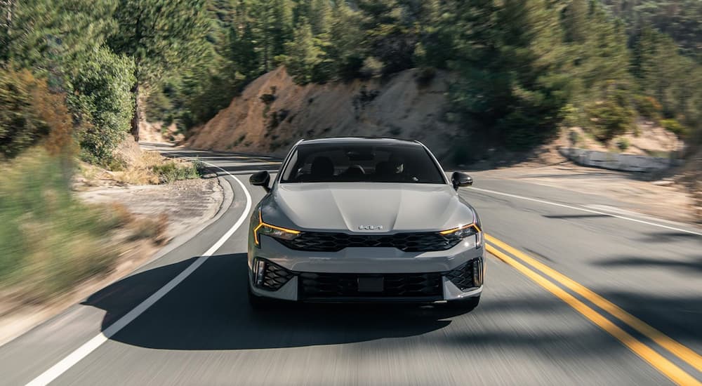 Front of a grey Kia K5 for sale driving down a road.