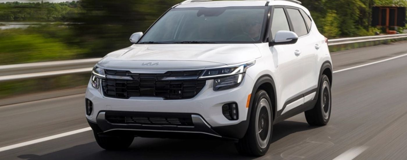 A white 2025 Kia Seltos is driving on an open road to a Kia dealer near Bloomington.