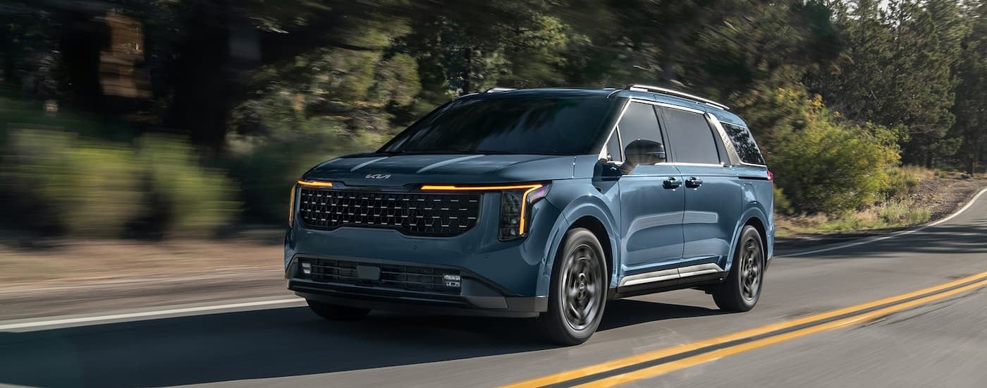 A blue 2025 Kia Carnival driving on a tree-lined road to a Kia dealer near Edina.