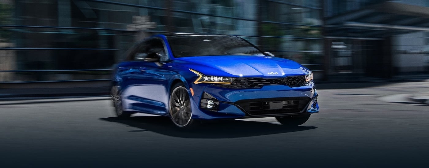 A blue 2024 Kia K5 driving in a city from a Kia Dealer fear St. Cloud